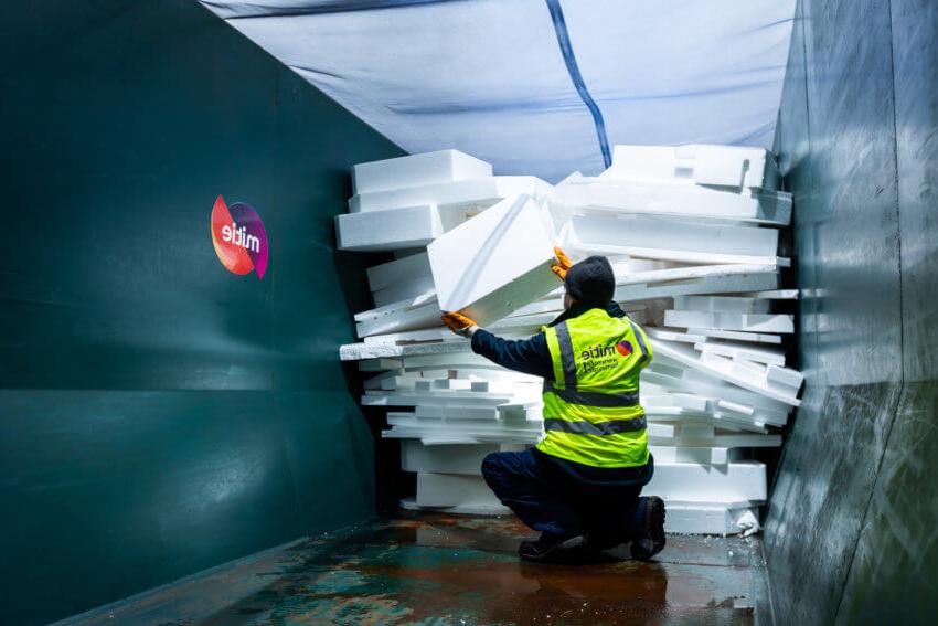 A man in a Mitie hi-vis vest sorting waste polystyrene in a dark green skip